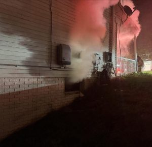 Smoke billows from an electrical panel on the exterior wall of a building at night, illuminated by red emergency lights, with a firefighter partially visible in the background.