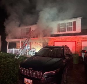 A two-story house with smoke pouring from the windows. A ladder leans against the front, and a black Jeep is parked in the driveway at night. Red emergency lights illuminate the scene.