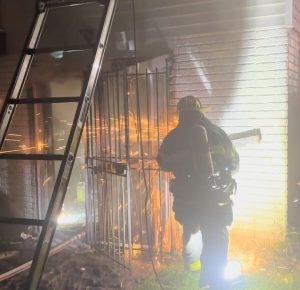 A firefighter in full gear uses a power tool to cut through metal bars on a building’s door at night, sending bright sparks flying, with a ladder leaning nearby against the structure.