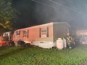 Firefighters work at night outside a single-story home with visible smoke and fire damage on one side. Fire equipment and a propane tank are nearby, and the scene is illuminated by emergency lights.