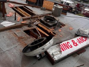 A damaged rooftop with large holes, debris, broken ventilation units, and a fallen sign reading “WING & ANC.” Insulation and wooden planks are exposed, and emergency vehicles are visible on the street below.