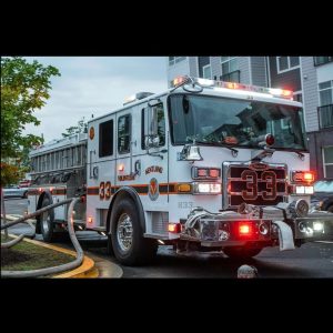 A white fire truck marked "33" and "VOLUNTEERS" is parked on a street next to an apartment building, with hoses connected and lights flashing, ready for emergency response.