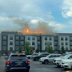 Flames and thick smoke rise from the roof of a four-story apartment building, while cars are parked in the lot in the foreground under a partly cloudy sky.