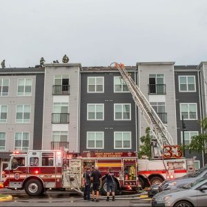 A firetruck with an extended ladder is parked in front of a modern apartment building. Firefighters are on the roof and several people stand near the truck, responding to an emergency.