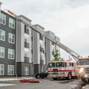 A fire truck with an extended ladder is parked outside a modern apartment building, reaching up to the roof. Emergency equipment lies on the ground nearby, and another vehicle is in the background.