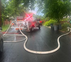 A fire department utility truck with flashing lights is parked on a wet residential street. Large fire hoses stretch from the truck, and trees and garbage bins line the road on a cloudy day.