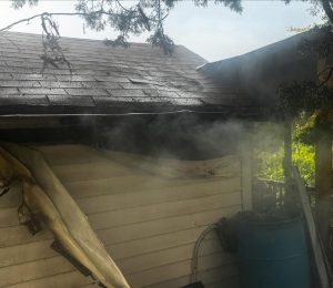 Smoke rises from under the roof of a light-colored building with visible fire damage. A partially melted tarp hangs over the side, and a blue barrel sits near the corner. Trees and sunlight are visible in the background.