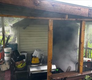 A covered porch with scattered household items, including buckets and a laundry basket, is filled with thick white smoke coming from a grill or smoker near the porch railing. The scene appears chaotic and cluttered.