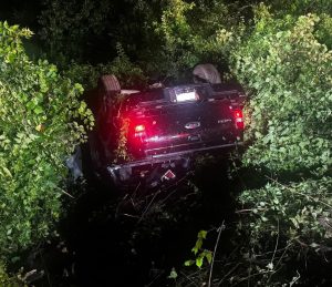 A black pickup truck flipped upside down in dense vegetation at night, with its taillights illuminated and partially obscured by branches and leaves.