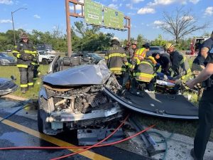 Firefighters work to rescue a person from a severely damaged car after a crash on a highway. The car's front end is crumpled, and its door has been removed. Emergency personnel are gathered around the scene.