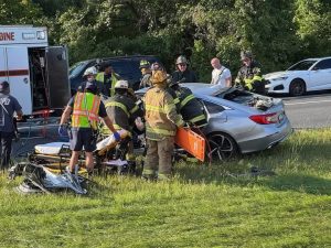 Firefighters and emergency responders assist a person on a stretcher next to a damaged silver car after an apparent accident, with emergency vehicles and other cars on the roadside in the background.