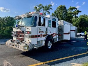 A white fire truck labeled "Kentland 33" is parked on a sunlit road with trees in the background. Two firefighters in reflective gear stand nearby, and the truck’s side compartment is open.