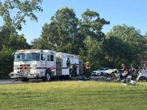 Firefighters and emergency responders work at the scene of a car accident involving severely damaged vehicles on the roadside, with a fire truck parked nearby and trees in the background under a clear sky.
