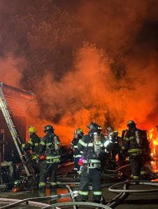 A group of firefighters in full gear works to control a large fire at night, surrounded by smoke, flames, hoses, and a ladder.