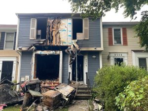 A townhouse with severe fire damage: the front doors and windows are missing or burned, exposing charred interiors and insulation. Debris and scorched items are scattered on the front lawn. The neighboring house is intact.