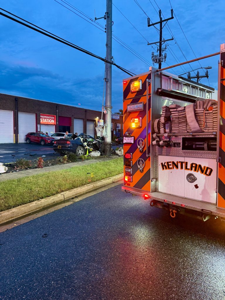 A fire truck with "Kentland" on the back is parked on a wet road near a car crash scene, where a black car appears to have collided with a utility pole outside a fire station at dusk.