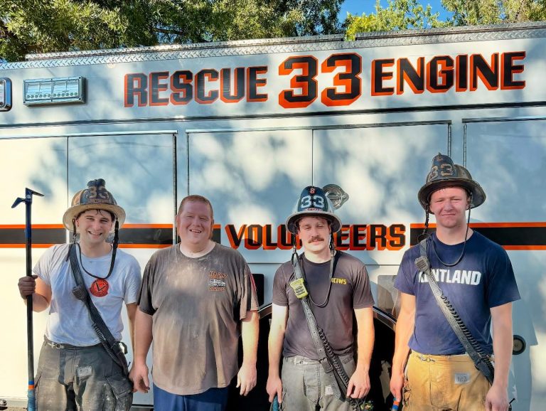 Four smiling firefighters in gear stand in front of a rescue truck labeled "RESCUE 33 ENGINE." Trees and blue sky are visible in the background.