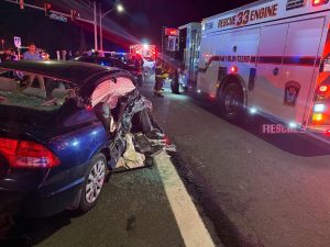 A dark car with severe rear-end damage sits on a road at night. Emergency vehicles, including a Rescue 33 fire engine, are present with flashing lights, and responders are at the scene.