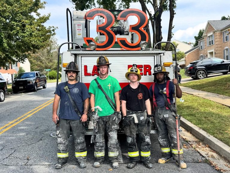 Four firefighters in gear stand in front of a fire truck with “33” on top, on a suburban street. They appear tired and covered in soot, holding equipment, with houses and trees in the background.
