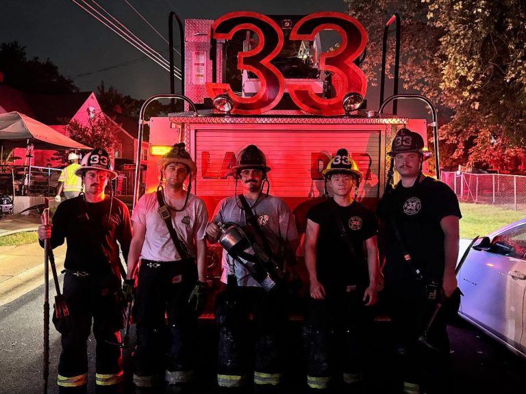 Five firefighters in gear stand in front of a fire truck with illuminated “33” on top at night, with red emergency lights casting a glow on the scene.
