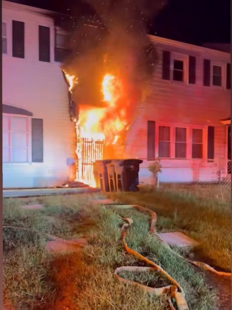 A large fire engulfs the exterior of a two-story house at night, with flames and smoke rising high. Fire hoses are stretched across the lawn, and two trash bins are near the blaze.