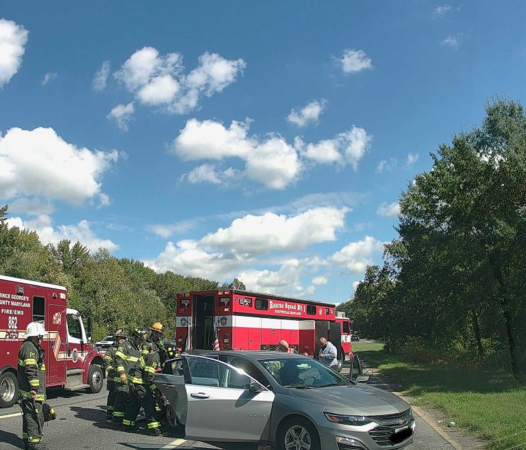 Firefighters and emergency personnel stand near a silver car involved in an accident on a road, with two fire trucks parked nearby and trees lining the scene under a blue sky with clouds.