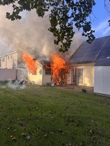 A yellow house with solar panels is engulfed in flames and thick smoke, especially around the front and roof. A bicycle is near the fire on the porch, and green grass is visible in the foreground.