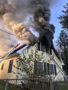 A yellow house with black smoke billowing out of upstairs windows and roof, indicating a fire inside. Ladders rest against the house, and a partial wooden fence is visible in the foreground.