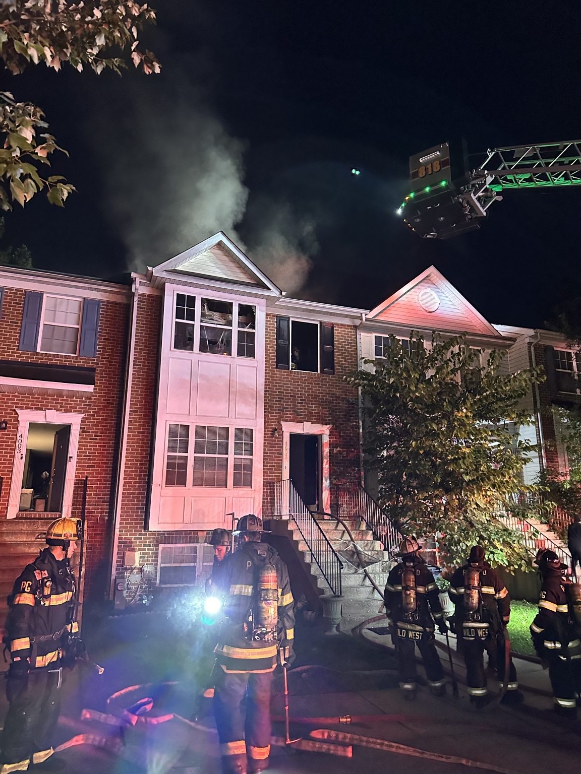 Firefighters stand outside a brick townhouse at night as smoke billows from upstairs windows. Emergency lights illuminate the scene, and a firetruck ladder extends toward the roof.