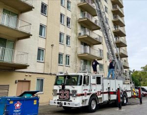 A fire truck with an extended ladder is parked next to a multi-story apartment building. Two firefighters stand on the truck near the ladder. A blue dumpster is visible in the foreground.