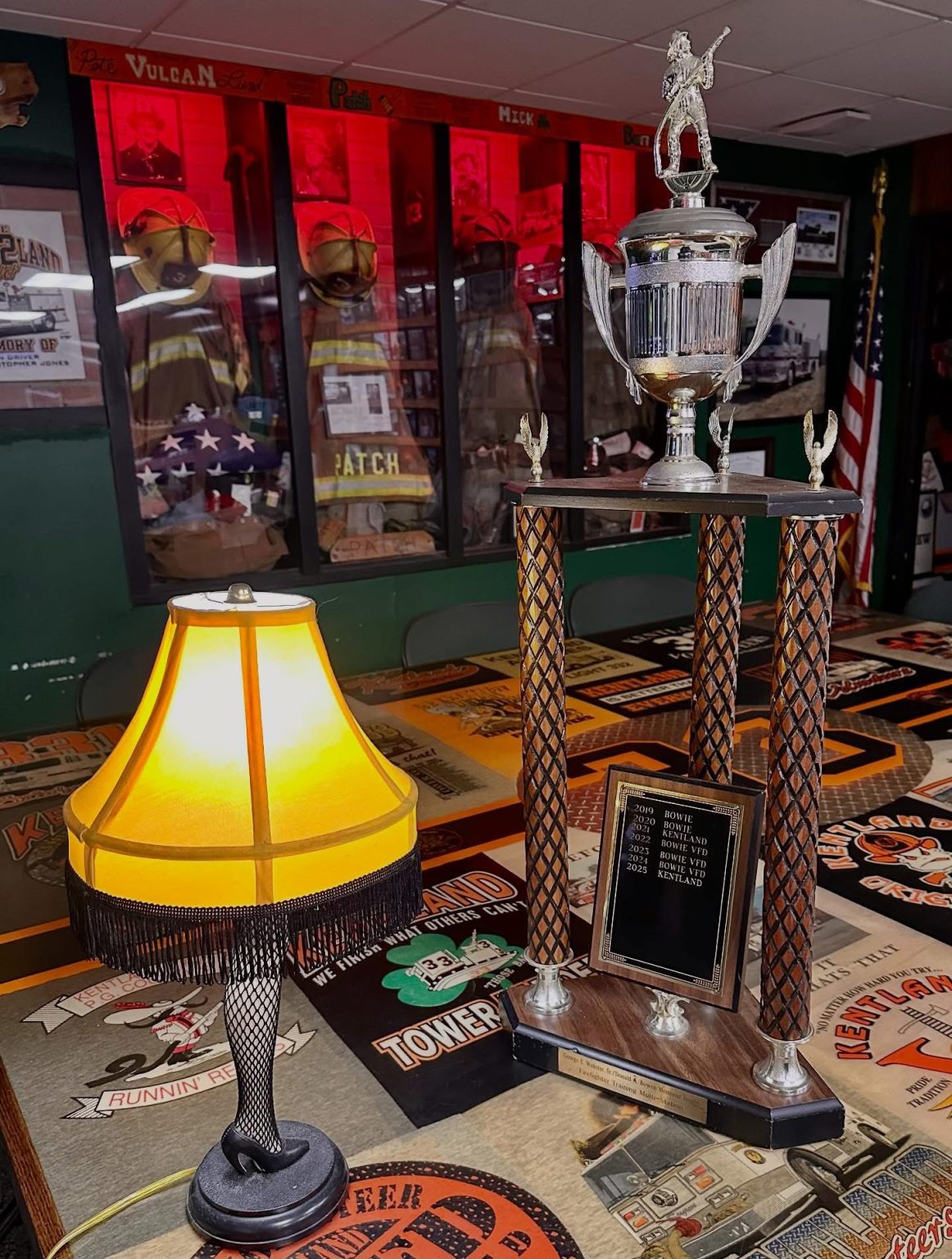 A leg-shaped lamp with a fringed shade sits on a table next to a large trophy. In the background are firefighter uniforms, helmets, and an American flag displayed behind glass.
