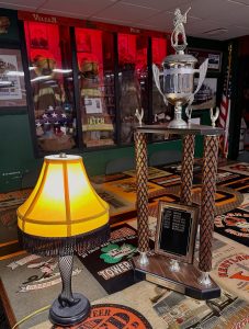 A leg-shaped lamp with a fringed shade sits on a table next to a large trophy. In the background are firefighter uniforms, helmets, and an American flag displayed behind glass.