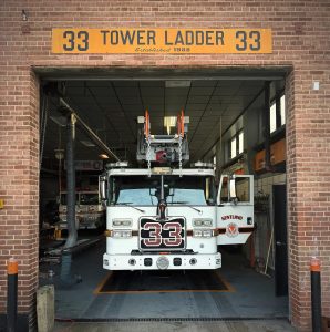 A white fire truck labeled "33 VENTLAND" is parked inside a brick fire station garage under a yellow sign that reads "33 TOWER LADDER 33 Established 1938." Equipment and tools are visible inside the station.