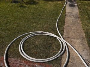 A coiled white hose lies on a grassy lawn near a brick edge, with another length of hose extending along a concrete sidewalk. A metal chair sits blurred in the background.