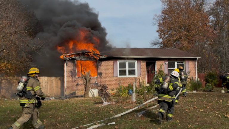 Firefighters approach a brick house with intense flames and thick black smoke pouring from a front window, preparing to extinguish the house fire. The yard has hoses and scattered debris.