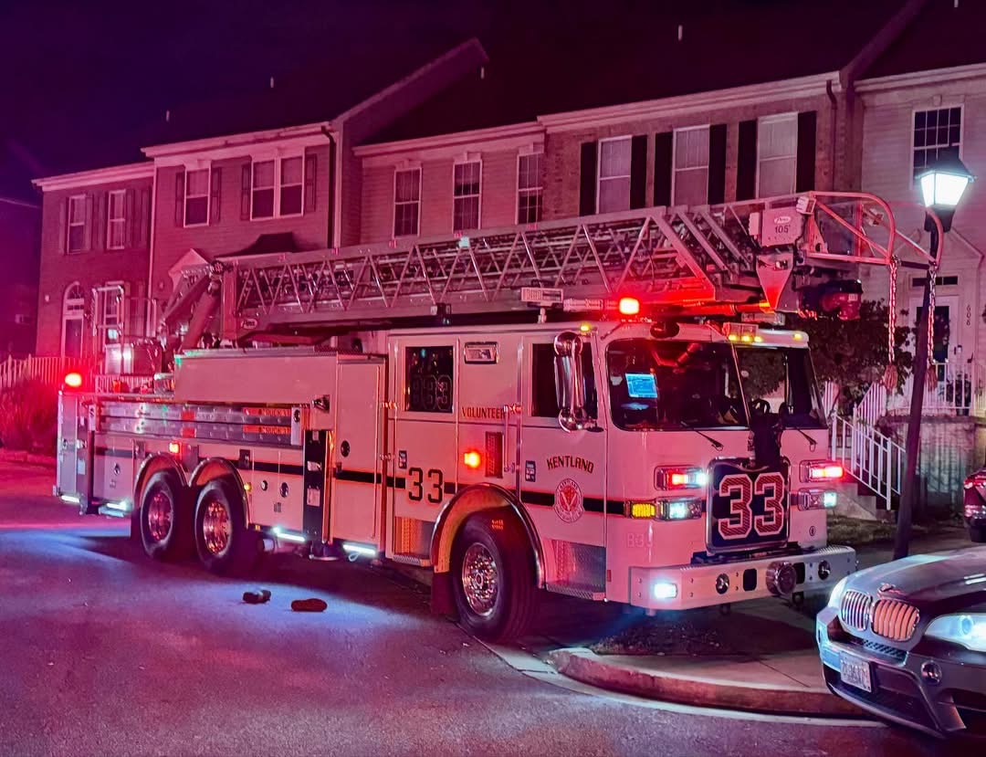 A fire truck with an extended ladder and flashing red lights is parked on a residential street at night in front of a row of townhouses. The number 33 is visible on the truck.
