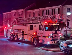 A fire truck with extended ladder and flashing lights is parked on a residential street at night in front of townhouses. The truck is labeled "33" and "VOLUNTEER." The scene is illuminated by emergency lights.
