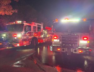 Two fire trucks with flashing lights are parked on a wet road at night. The scene is illuminated by the headlights and emergency lights, with reflections visible on the pavement. Trees are partially visible in the background.