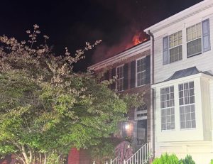Flames are visible on the roof of a two-story brick townhouse at night. A tree partially obscures the view, and smoke rises from the top of the building. The house next door is not affected.