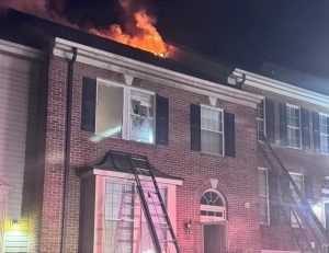 Flames and smoke rise from the roof of a brick townhouse at night. Broken windows and ladders leaning against the building suggest emergency response activity.
