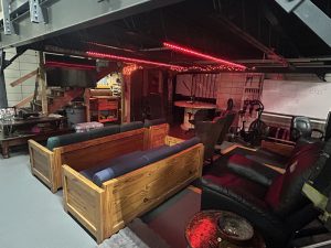 A cozy basement lounge features wooden couches, black recliners, a table, and red ceiling lights. Exposed stairs and a neon sign are visible, with eclectic decor and a whiteboard in the background.