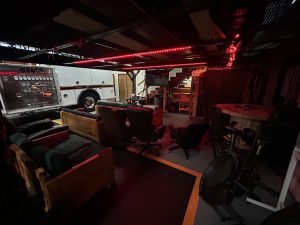 A dimly lit room features several chairs, wooden benches, a round table, and TV, with red LED lights overhead. A large fire truck is parked inside, and stairs lead up to a loft area.