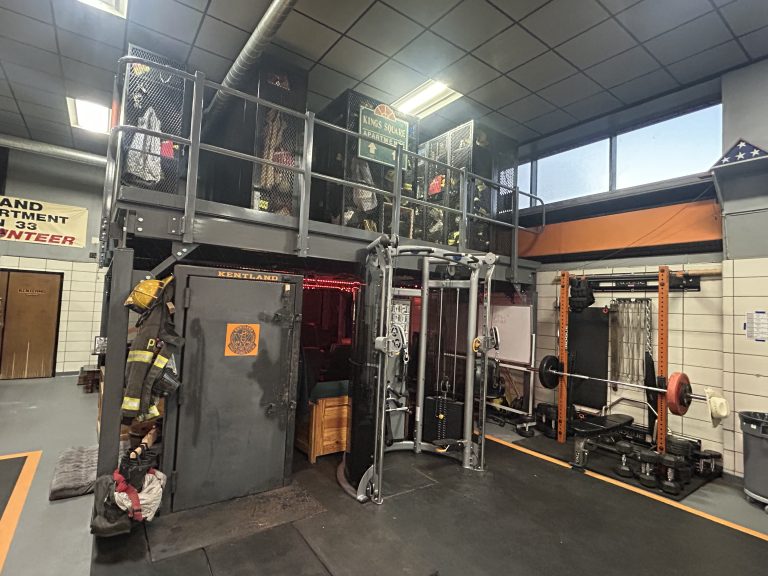 A gym with weightlifting equipment, a power rack, and barbells. Above the gym area is a storage loft with firefighter gear and cages. Walls are decorated with firefighter memorabilia and American flag.