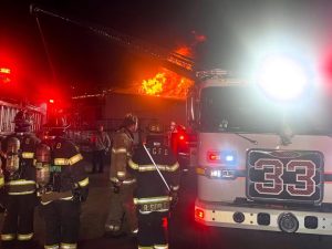Firefighters stand near a firetruck labeled “33” at night, responding to a large building fire with intense flames and smoke rising in the background. Red emergency lights illuminate the scene.