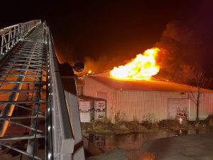 A large fire blazes through the roof of a metal building at night, with flames and smoke rising. A fire truck’s ladder is extended toward the building in the foreground.