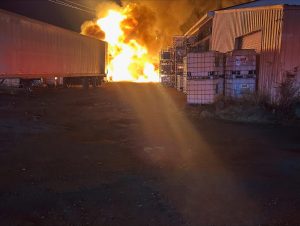 A large fire blazes at night between an industrial building and a parked trailer, with flames lighting up the sky and industrial containers stacked nearby.