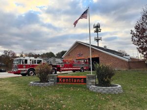 A red fire truck is parked outside a brick fire station with an American flag flying on a pole. In front, a sign reads "Kentland" surrounded by small shrubs and a stone border. The sky is cloudy.