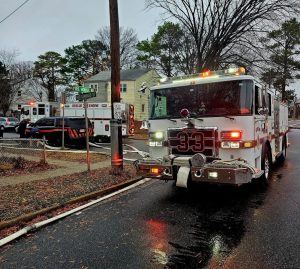 A fire truck with the number 33 is parked on a wet residential street near a corner with a rescue vehicle and an ambulance. Houses, leafless trees, and emergency personnel are visible in the background.