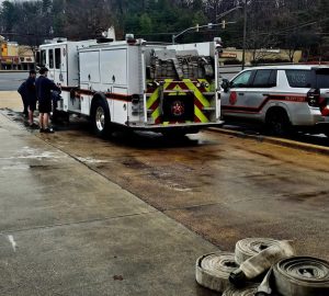 A fire truck and a fire department SUV are parked on a wet pavement near several rolled fire hoses. Two people stand by the fire truck, and a few buildings and trees are visible in the background.