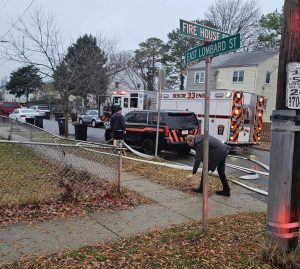 A man crouches on a sidewalk near a street sign for "Fire House St" and "East Lombard St" as fire trucks and a rescue vehicle with flashing lights are parked on the street in a residential neighborhood.
